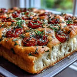 Fluffy Italian focaccia bread scattered with vibrant spring herbs, ripe cherry tomatoes, and a sprinkle of flaky sea salt.  